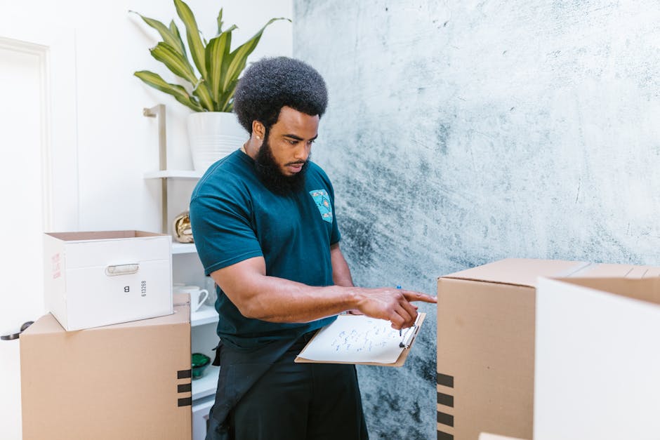 A young male mover, wearing a dark blue work uniform and a black bandana on his head, is standing inside a room with a textured blue wall background. He is holding a clipboard and pen, appearing to ta