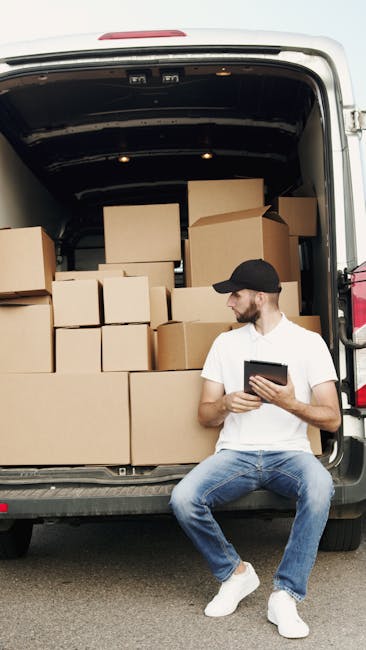 A man wearing a white t-shirt, black cap, and jeans is sitting on the ground in front of a high-sided van with an open rear door, which is filled with stacked cardboard boxes of various sizes. Some bo