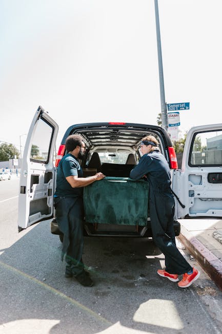 A man wearing a white t-shirt, black cap, and jeans is sitting on the ground in front of a high-sided van with an open rear door, which is filled with stacked cardboard boxes of various sizes. Some bo