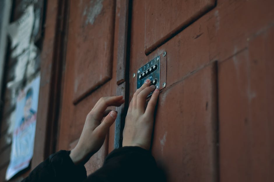 A person's hands are seen operating a combination lock on a wooden door, with one hand pressing a dial while the other stabilizes the lock. The door is made of reddish-brown timber with visible grain and paneling, and it has a metal lock mechanism with numbered dials. To the left, part of a metal handle is visible, and the door appears to be indoors or at the entrance of a house. The background shows blurred elements, possibly a corridor or porch area, and soft lighting highlights the texture of the wood and the metallic lock. This image reflects the process of securing a property or preparing for a home relocation, as performed by [COMPANY_NAME], who assist with house moving services, especially in situations involving narrow access homes. The focus on the lock and door emphasizes key aspects of logistics and security during furniture transport and packing in the context of house removals.