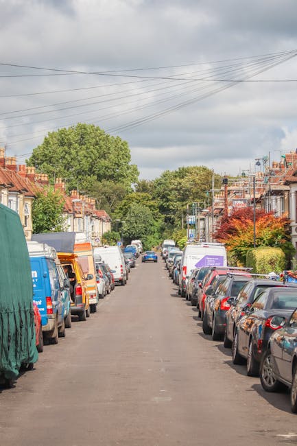 A residential street with closely spaced terraced houses and well-maintained front gardens, lined with a variety of parked vehicles including vans, cars, and moving trucks on both sides of the narrow asphalt road. Some vans are covered with protective fabric or plastic wrapping, indicating active packing or wrapping for a home relocation process. The scene shows a typical urban neighbourhood environment with overhead power lines, trees, and a cloudy sky overhead. This setting illustrates a logistical challenge often encountered during house removals, especially in streets with restricted access. The image captures a period of active furniture transport and loading operations, with vehicles positioned near house entrances, reflecting a professional move managed by companies like Highbury Removals, which specialise in addressing narrow access homes requiring detailed planning for effective packing, loading, and transport.