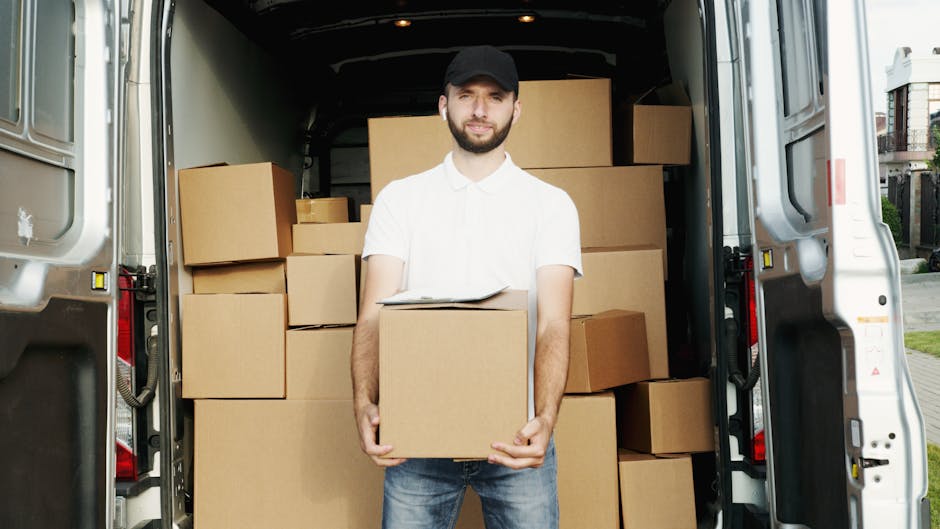 A young man wearing a white polo shirt and a black cap stands inside the open back of a grey panel van, holding a cardboard box during a house relocation. The van is fully loaded with several other cardboard boxes of different sizes, some stacked on top of each other and others arranged along the sides. The boxes appear to be secured for transport and are made of brown corrugated cardboard, with some wrapped in plastic or packing materials. The interior of the van is spacious, with a black ceiling and walls, and the boxes are positioned adjacent to the vehicle's interior walls, ready for loading or unloading. The scene takes place outdoors, on a paved area, possibly a driveway or street near a residential property, with some greenery and buildings visible in the background. This image illustrates the process of furniture transport, packing, and loading during professional home relocation services delivered by Highbury Removals, with an emphasis on careful handling and organized moving logistics.