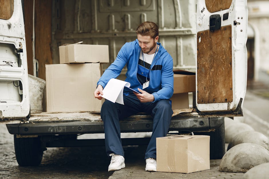 A man wearing a black and grey baseball cap, a black quilted vest over a blue long-sleeve shirt, and black gloves, stands next to an open white moving van during a house relocation. Inside the van, visible are several cardboard boxes, some wrapped in plastic film, and protective blankets covering furniture. The man appears to be preparing or inspecting items for loading, with his hands close together. The background shows a brick exterior wall of a residential property with a grey fence and greenery, suggesting an outdoor loading area on pavement. The scene illustrates the process of furniture transport and packing in a professional house removal service, with Highbury Removals subtly indicated by the context.